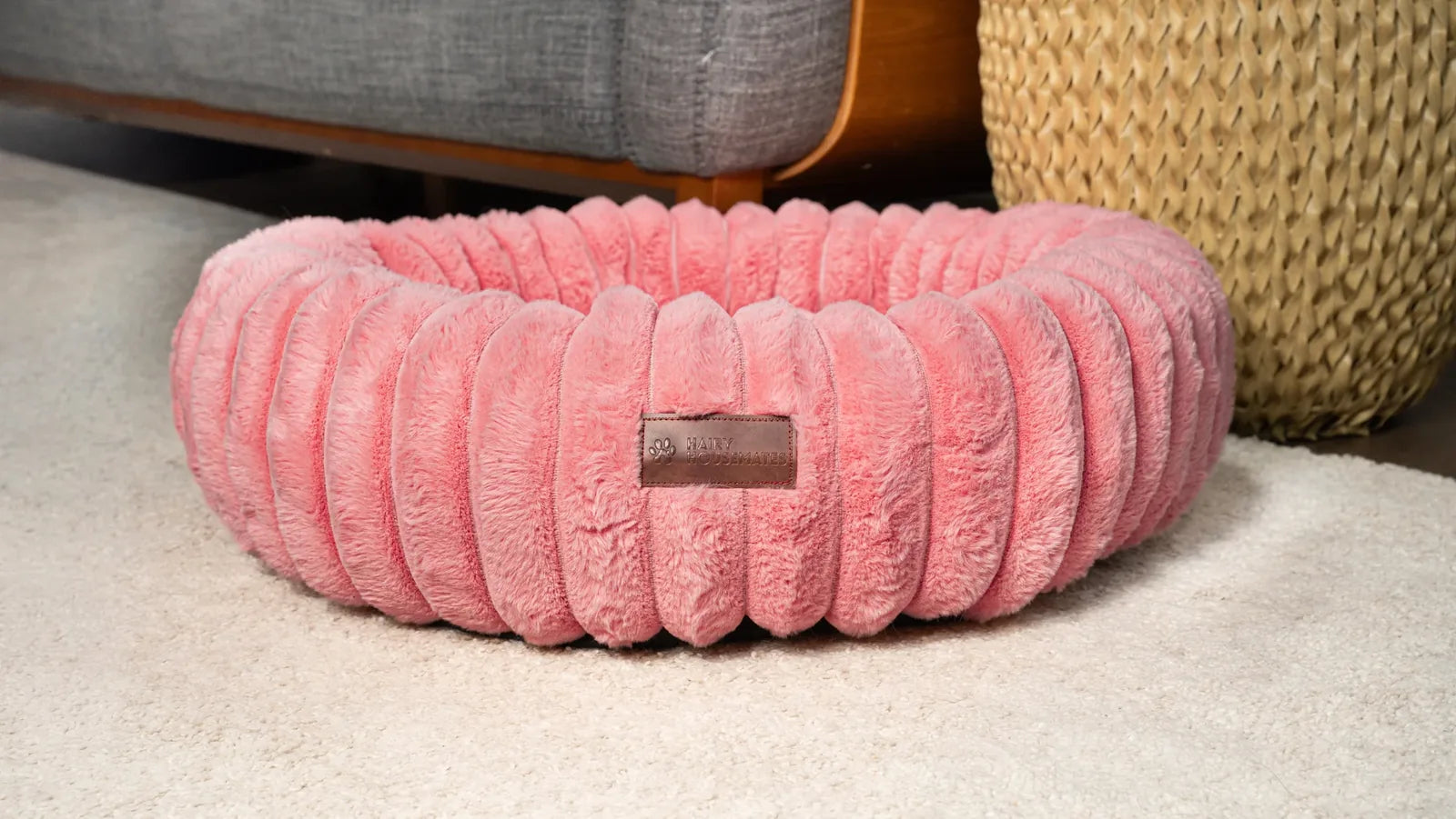 A pink corduroy dog bed styled in a bright room corner beside a sofa